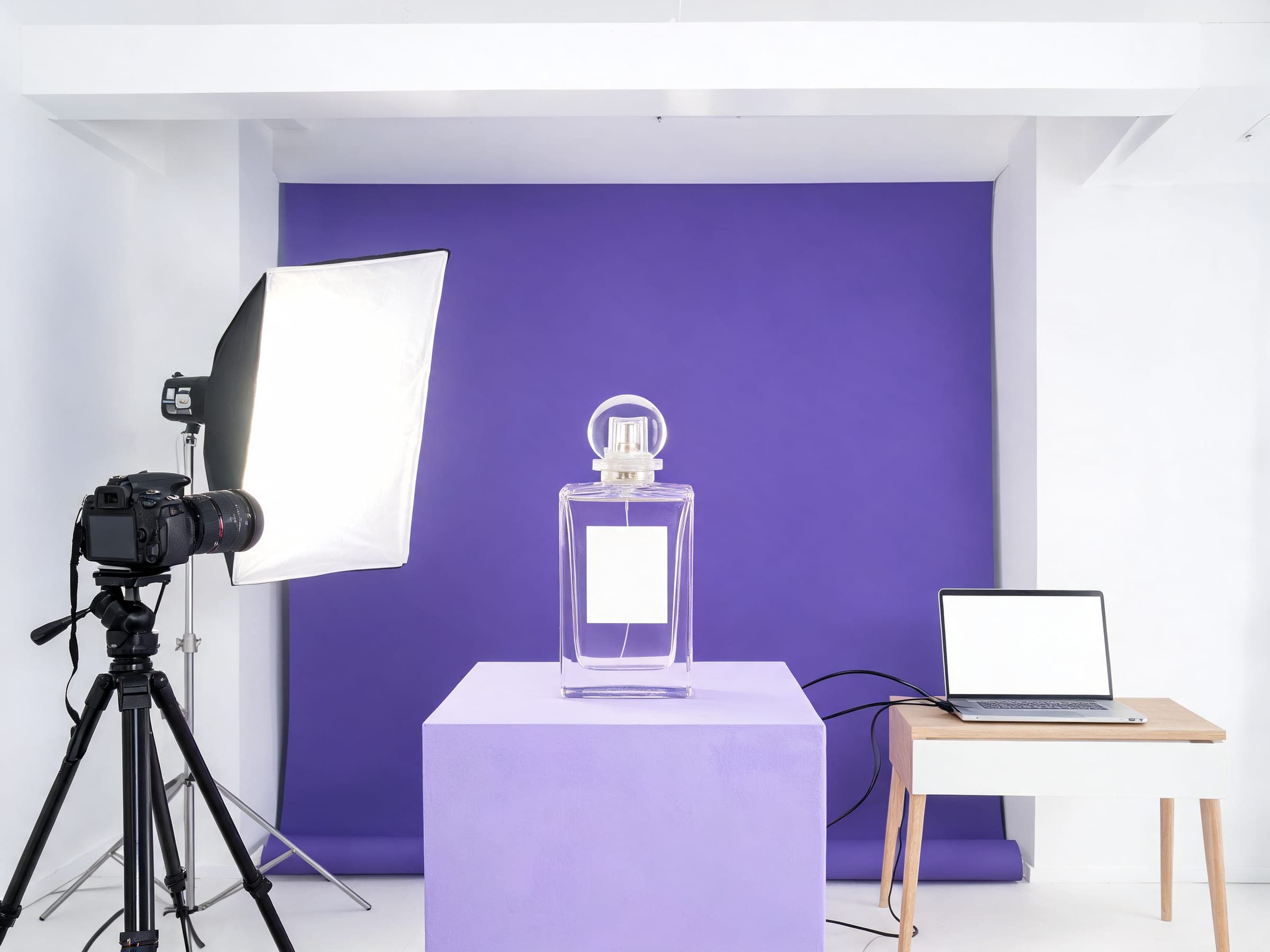 A small studio set up to shoot a perfume bottle against a violet backdrop, with a camera, a softbox and a laptop