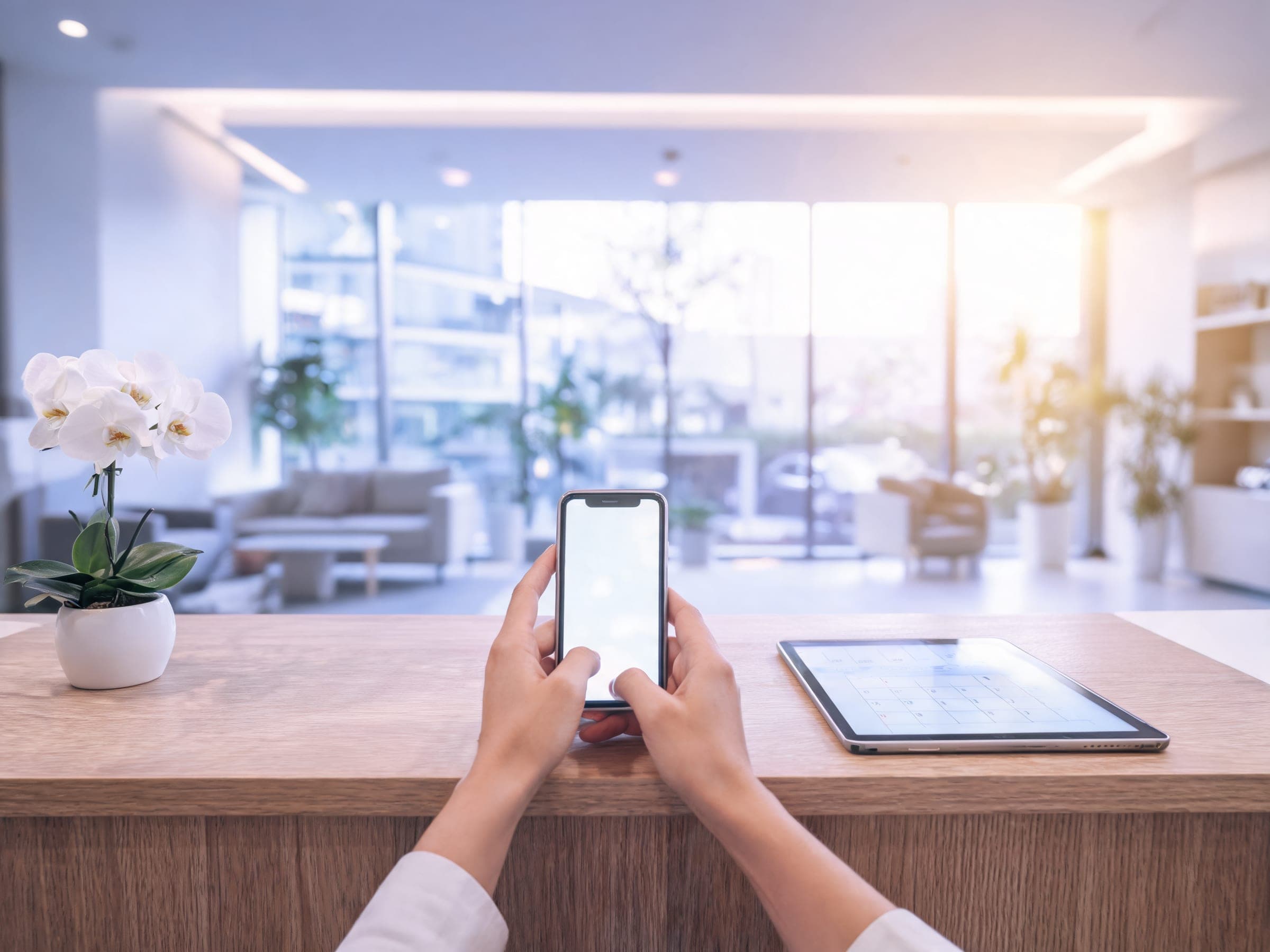 Hands holding a phone at a clinic’s front desk, a tablet calendar beside it
