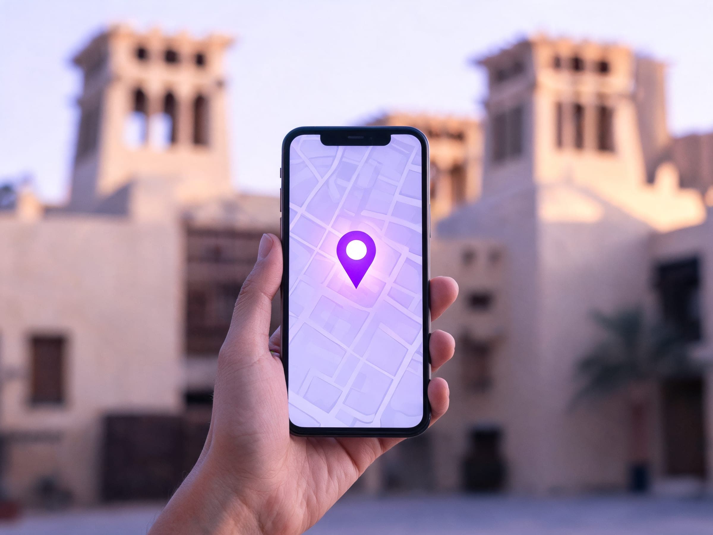 A hand holding a phone with a map pin, in front of the wind towers of Al Fahidi in Dubai