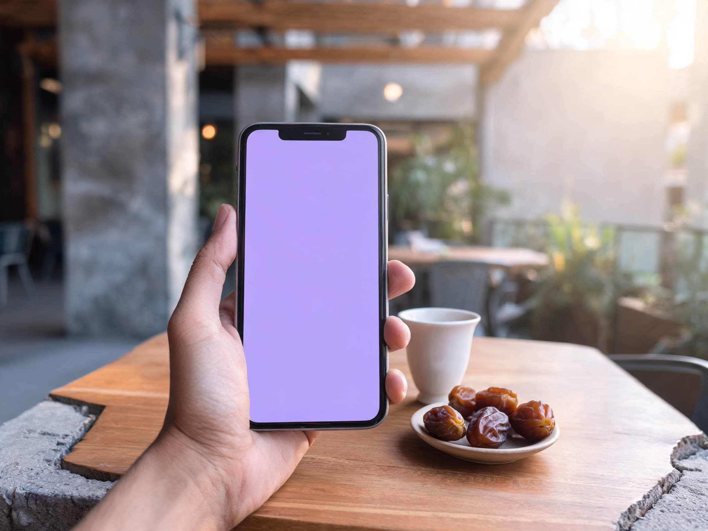 A hand holding a phone at a sunlit Dubai café table with coffee and dates