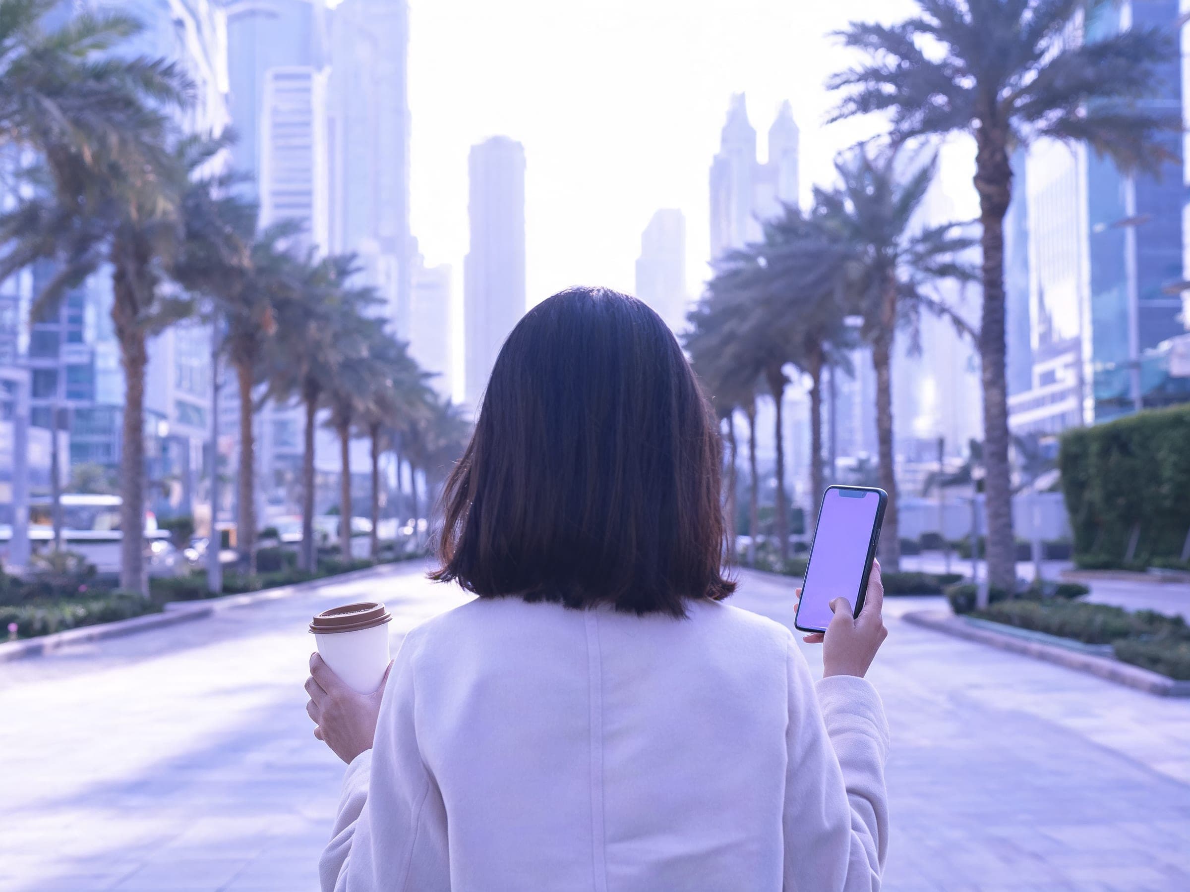 Seen from behind, a person with a coffee checks a phone on a palm-lined street in a Dubai business district